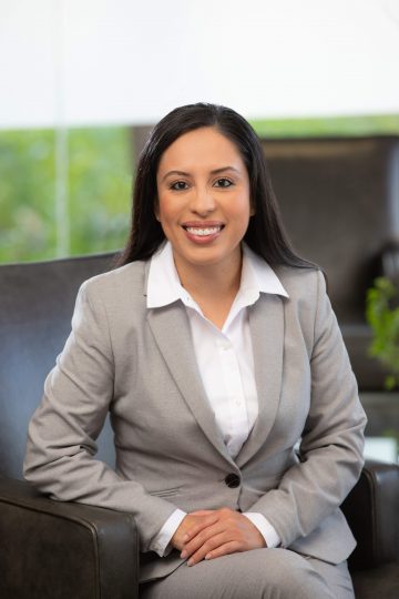 A hispanic lawyer wearing a gray suit, sitting in a chair with her hands folded.