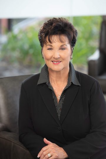 A woman with short brown hair in a gray suit seated in a brown chair.