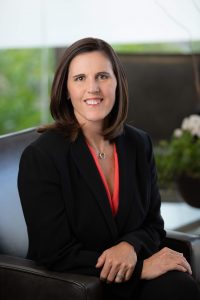 A woman with shoulder-length brown hair in a black suit, seated in a brown chair with her arms crossed